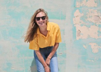 Casual young woman smiling in sunglasses, posing against a vintage colorful wall.