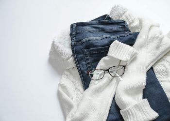 Stylish flat lay featuring denim jeans, white sweater, and eyeglasses on a bright background.